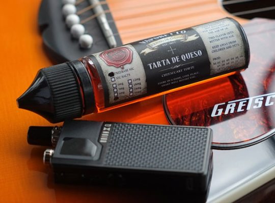 a black vape device and a cheesecake flavored liquid cartridge lying on a guitar