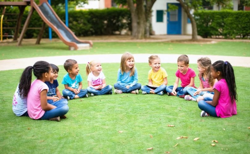 A group of children of diverse backgrounds sitting in a semicircle on a grassy field, actively engaged in storytelling and practicing communication and active listening skills with the guidance of an adult.