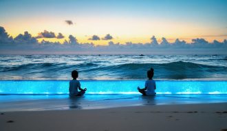 Silhouettes of children practicing meditation at sunrise on a beach, surrounded by gentle blue ocean waves with a subtle blue glow, symbolizing the calming effect of blue light meditation.