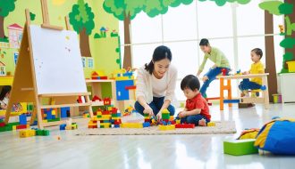 A kindergarten playroom filled with natural light, showcasing various activity zones with children and parents engaged in play-based learning. A parent and child smiling as they play with building blocks, illustrating family bonding through play.