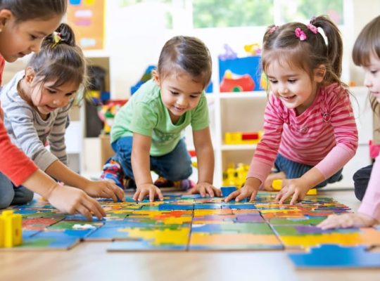 A diverse group of children happily collaborating on assembling a large puzzle on the floor, demonstrating teamwork and communication in a playful setting.